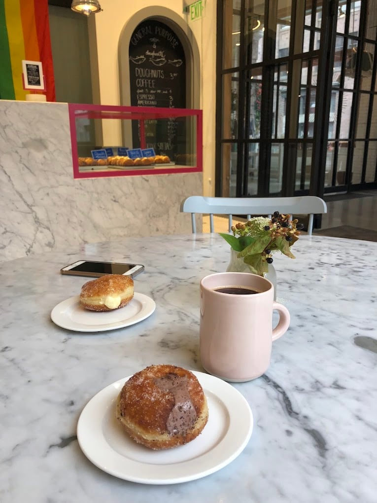 A marble table with two plates of filled doughnuts, a mug of coffee, a phone, and a small vase with greens. In the background, a counter displays pastries and a chalkboard menu beneath a pride flag.
