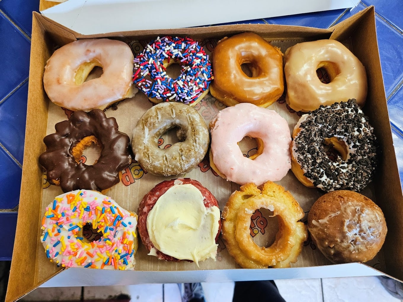 A box of twelve assorted donuts, including glazed, chocolate, pink icing, sprinkles, cookies and cream, and cake donuts, arranged in three rows on Dunkin' branded paper.