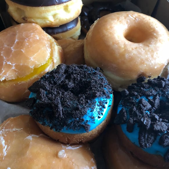 A close-up of assorted donuts, including glazed, chocolate-covered, jelly-filled, and donuts topped with bright blue icing and crumbled cookies, arranged closely together in a box.