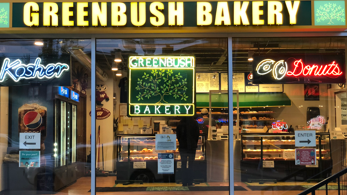 Storefront of Greenbush Bakery with bright yellow sign, neon "Kosher," "Greenbush Bakery," and "Donuts" signs, and display cases filled with baked goods. Doors marked "Exit" and "Enter" with arrows.
