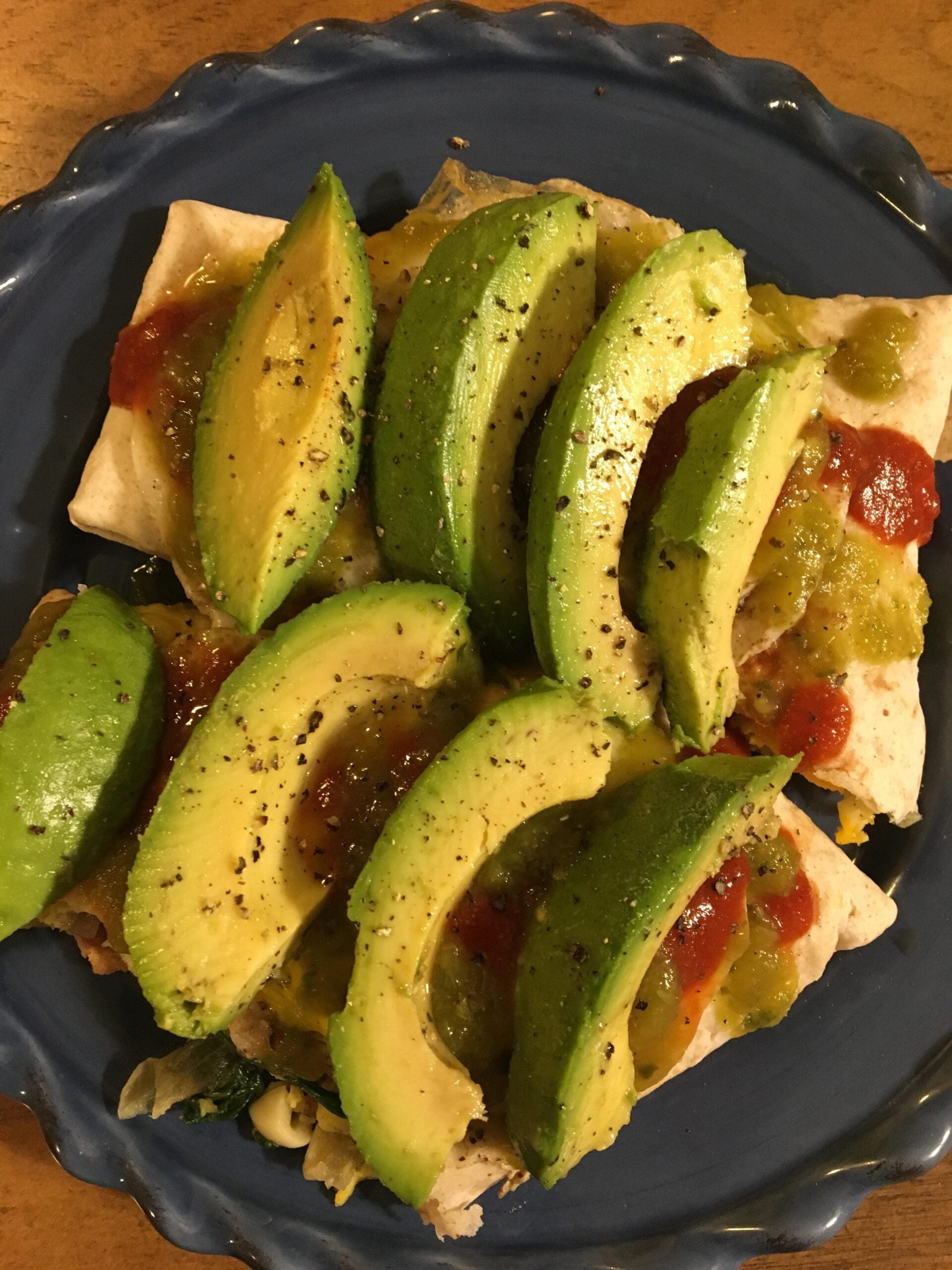 A blue plate with burritos topped with sliced avocado, salsa, green sauce, and black pepper.
