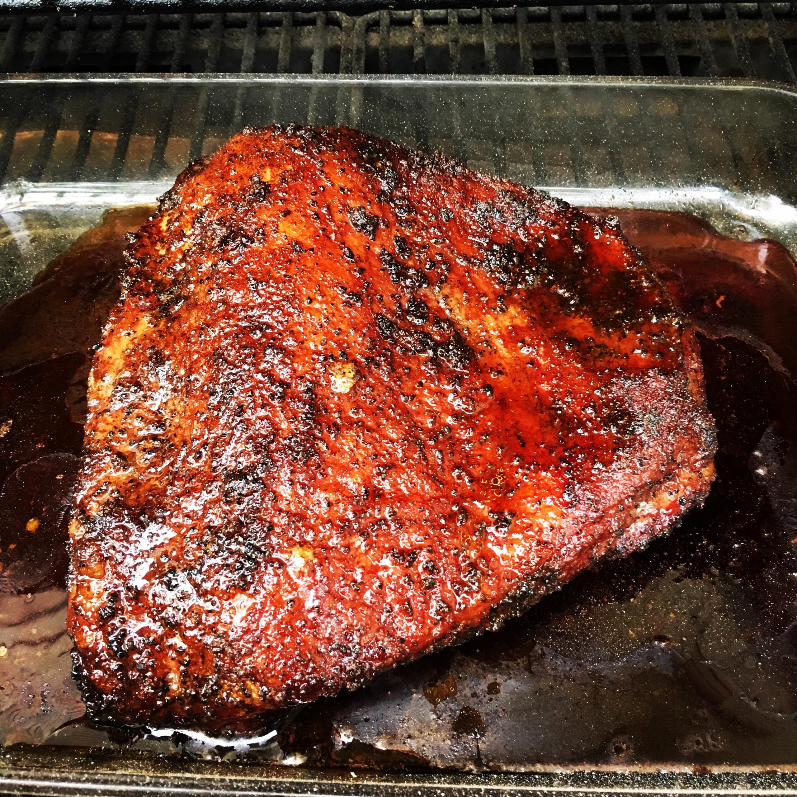 A large, juicy, smoked brisket with a dark, flavorful crust sits in a glass pan on a grill, glistening with juices and charred seasoning.