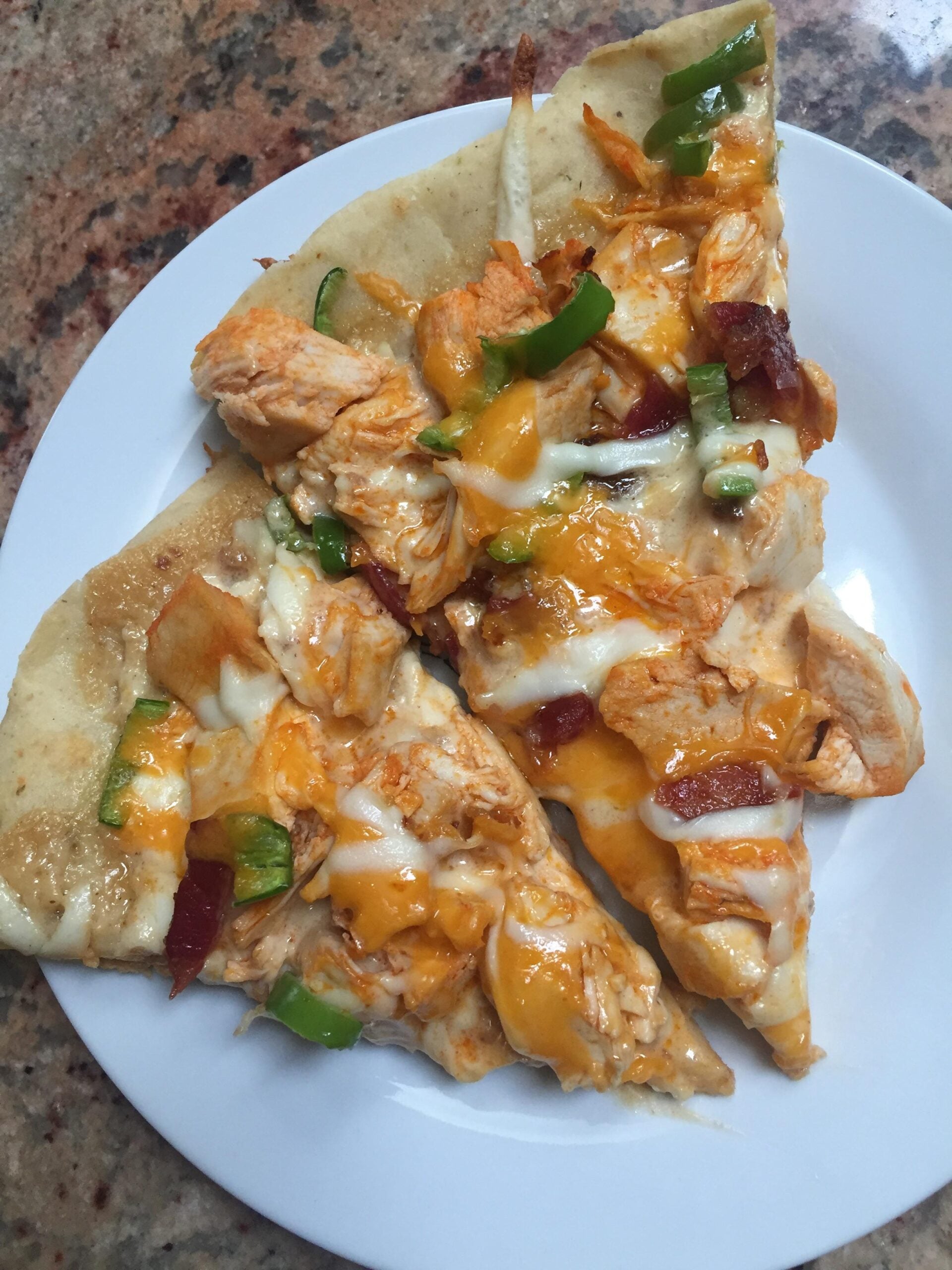 Three slices of pizza with melted cheese, chicken pieces, green bell peppers, and bacon bits on a white plate, set on a speckled countertop.