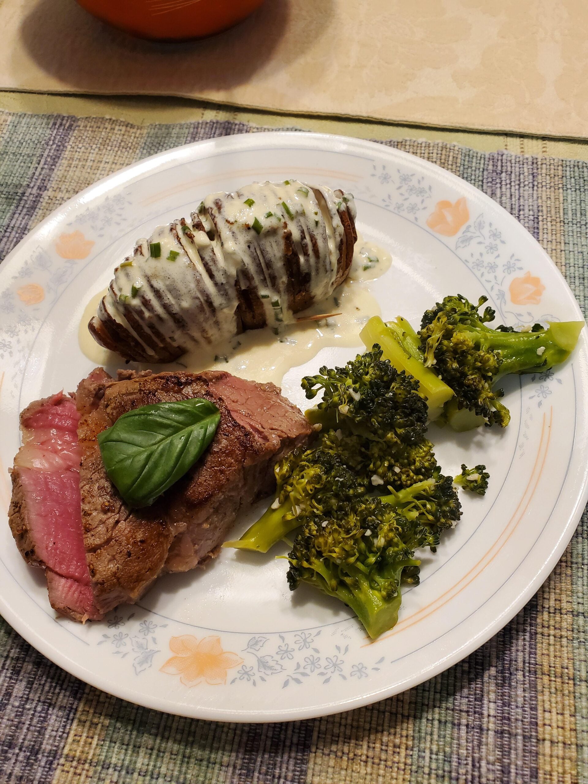 A plate with sliced steak topped with a basil leaf, roasted broccoli, and a hasselback potato drizzled with creamy sauce and chives, on a patterned plate set on a woven placemat.