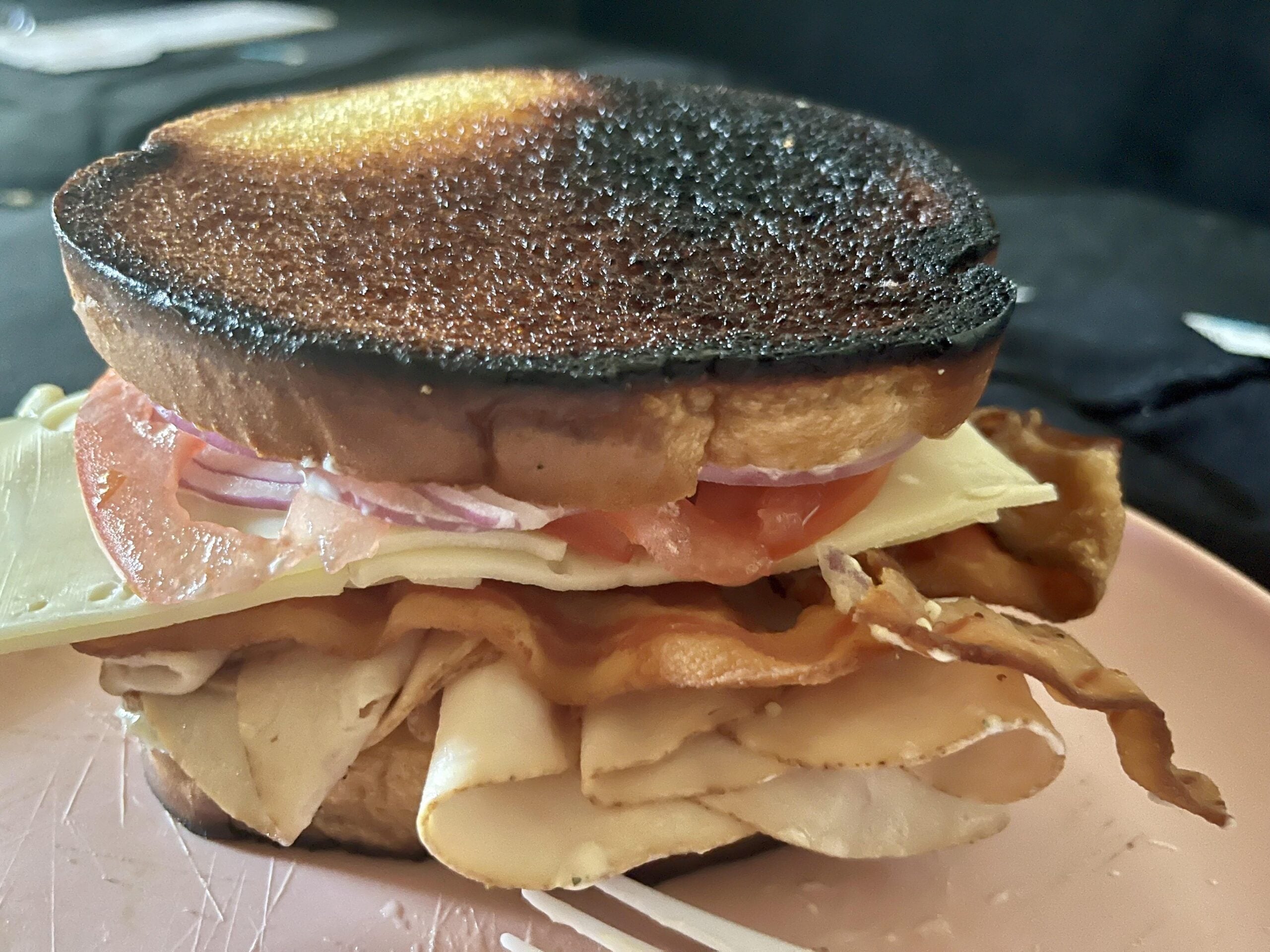 A sandwich with burnt toast, turkey, bacon, cheese, tomato, and red onion sits on a pink plate.