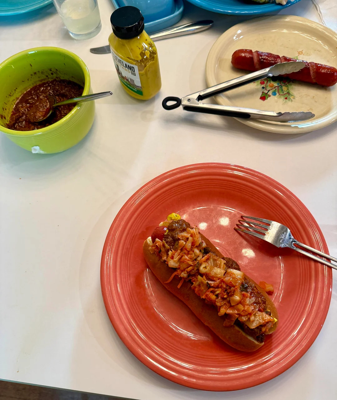 A chili dog topped with onions and mustard on a coral plate, a fork beside it. Nearby are a plain hot dog, bowl of chili, mustard bottle, and kitchen tongs on a white table.