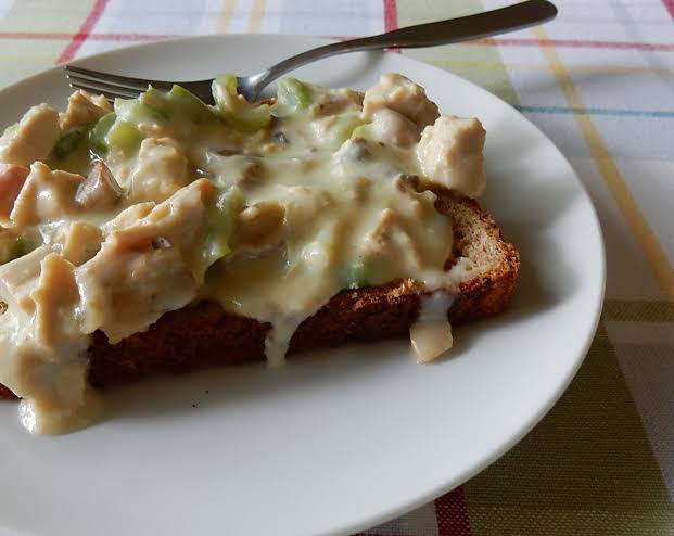 1950s family meals. A slice of bread topped with creamy chicken and vegetable mixture is on a white plate with a fork. The plate sits on a checkered tablecloth.