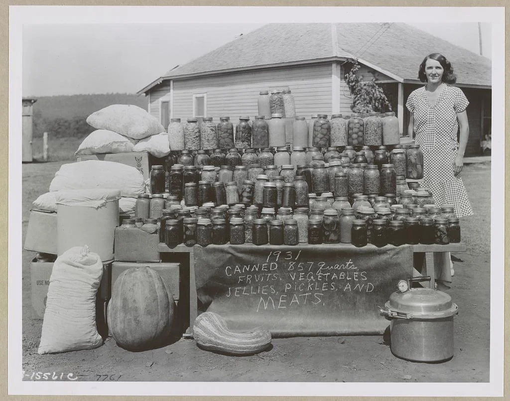 A woman stands beside a display of canned goods, sacks, and produce in front of a house. The sign reads “1931 canned 857 quarts fruits, vegetables, jellies, pickles, and meats.”