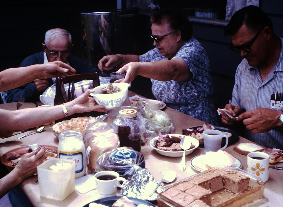 Four adults sit around a table sharing a meal with various dishes, desserts, coffee, and bread. One woman serves food, while others reach for items. The scene appears casual and warm, set outdoors or on a porch.