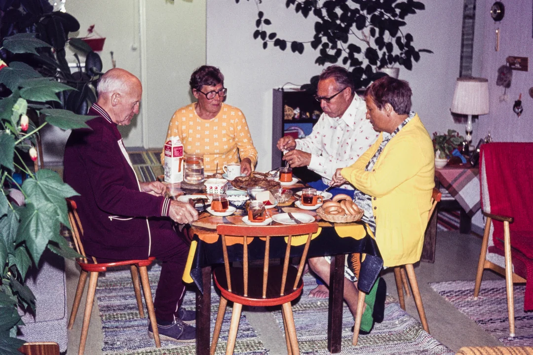 Four older adults sit around a small table enjoying coffee and pastries in a cozy, plant-filled living room. They appear relaxed and engaged in conversation.
