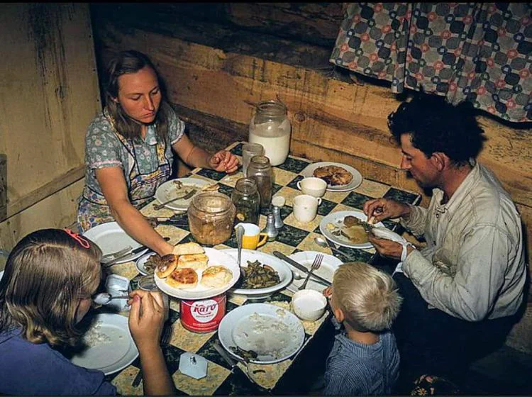 A family of four sits around a table in a rustic wooden room, eating a meal together. The table holds plates of food, jars, a pitcher of milk, and a container labeled “Fairy.”