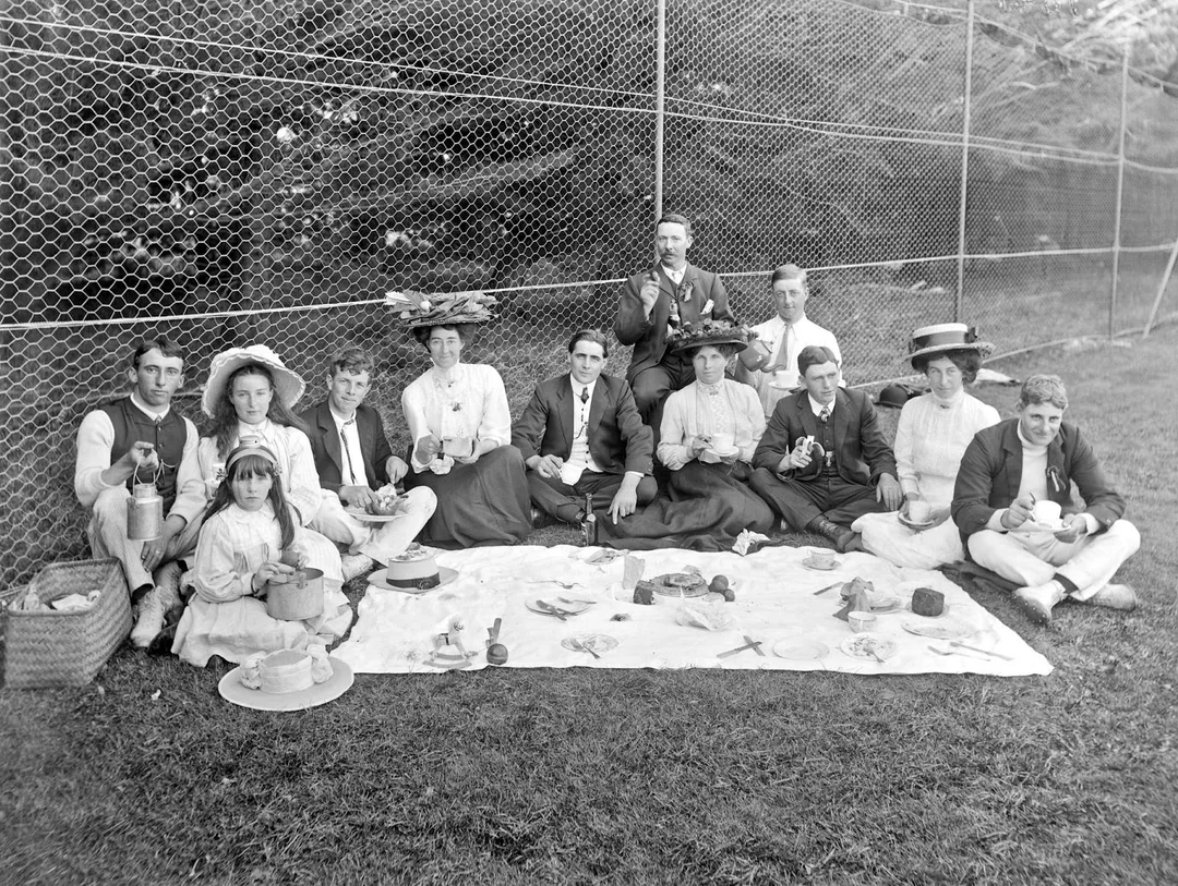 Thirteen people in vintage clothing have a picnic on a lawn. They sit on a blanket with tea cups and food, posing for the camera. There’s a chain-link fence and trees in the background.