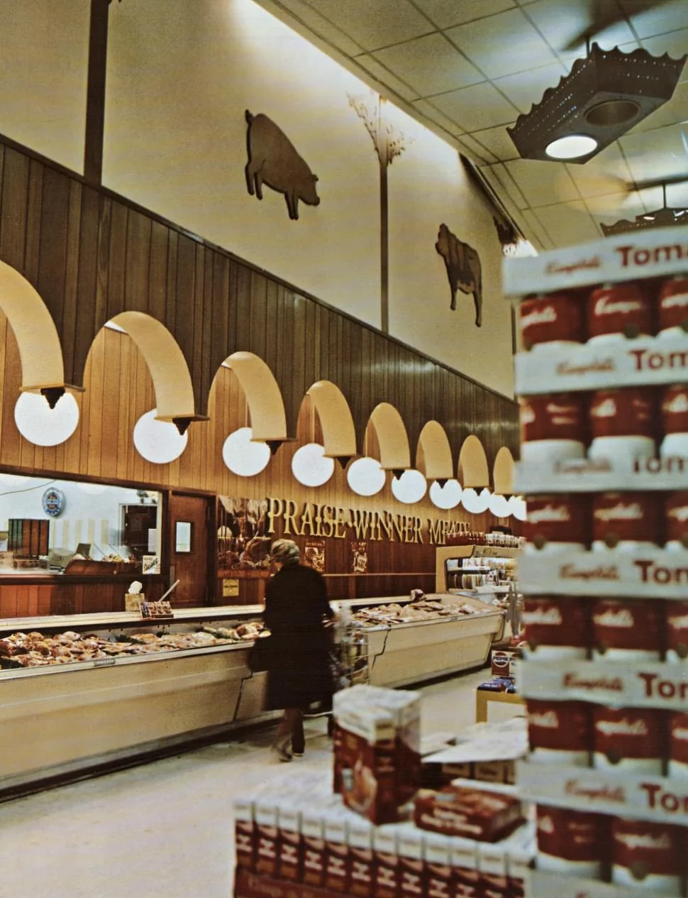 A mid-century grocery store aisle featuring a meat counter labeled "Praise Winner Meats." Above are wood-paneled walls with pig and cow silhouettes and arched lamps. In the foreground, a shopper walks near a large stack of Campbell's soup cans.