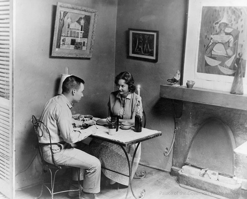 Black-and-white historical photo of a man and a woman sitting at a small table for a candlelit meal. They are in a room decorated with artwork and a fireplace. The woman looks at the man as they eat. Palace of the Governors Photo Archives.