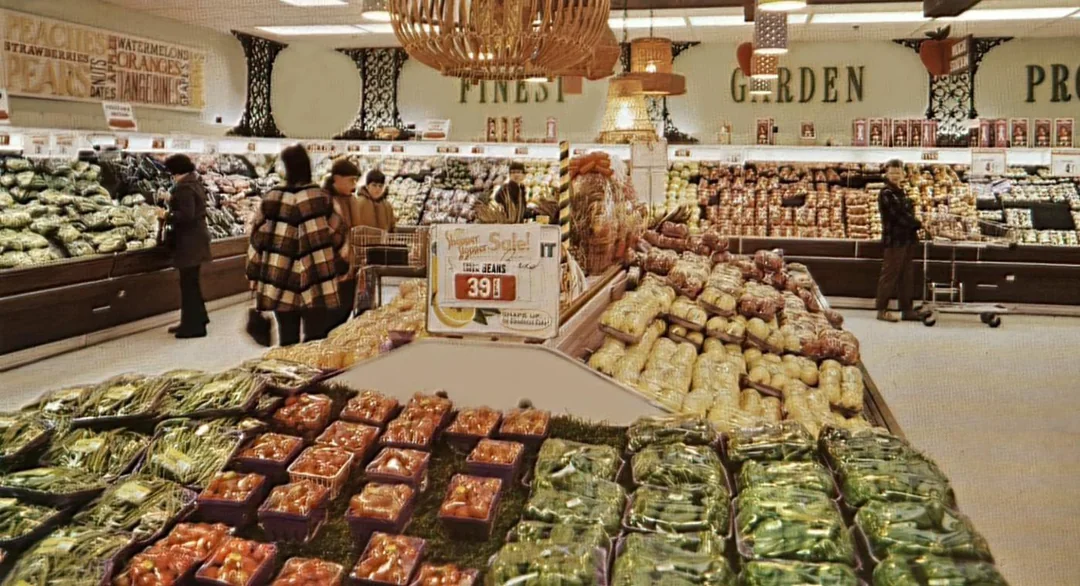 Shoppers browse a brightly lit supermarket produce section filled with fresh vegetables on display, such as peppers and tomatoes, with signs and baskets hanging above.