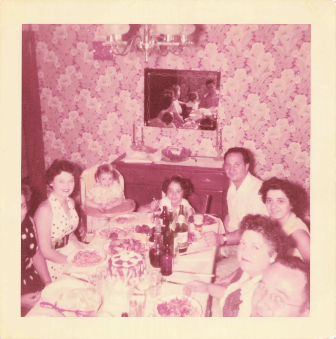 Six adults and two children sit around a dinner table covered with food and drinks, in a room with floral wallpaper and a mirror reflecting their image. The atmosphere appears warm and festive.