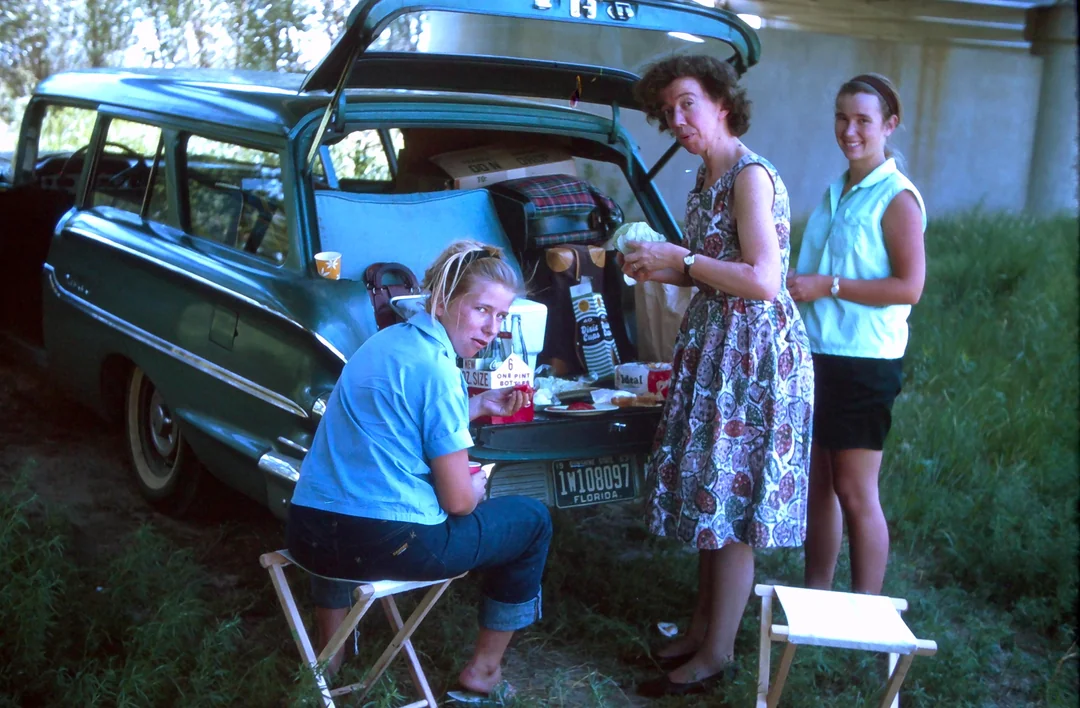 Three women prepare food at the open trunk of a green vintage station wagon, with stools and picnic items around them, outdoors on a sunny day.