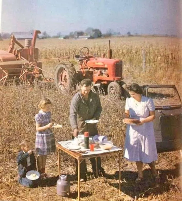 Vintage food photos. A family eats a meal at a small table in a field, with a red tractor and crops in the background. The mother, father, and two children are dressed in simple, old-fashioned clothing.