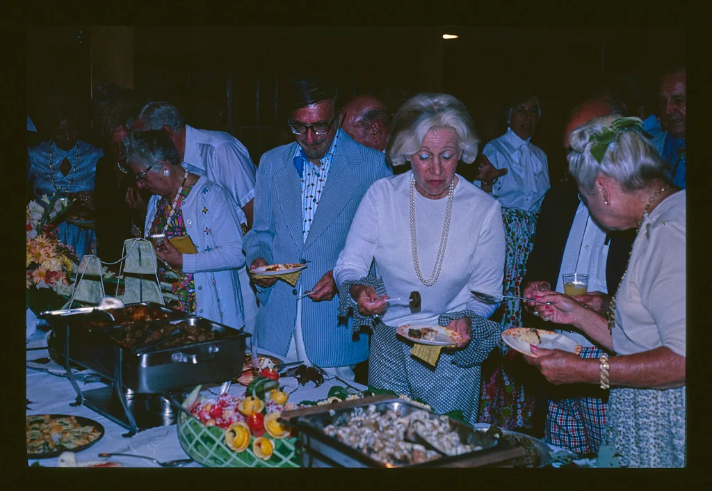 A group of older adults, dressed semi-formally, serve themselves food from a buffet table covered with various dishes at a social gathering.