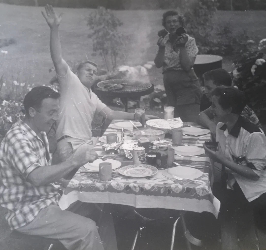 Four people sit at an outdoor table eating, while one stands behind, taking a photo. The table is covered with plates, cups, and food. One person raises an arm excitedly. Trees and plants are visible in the background.