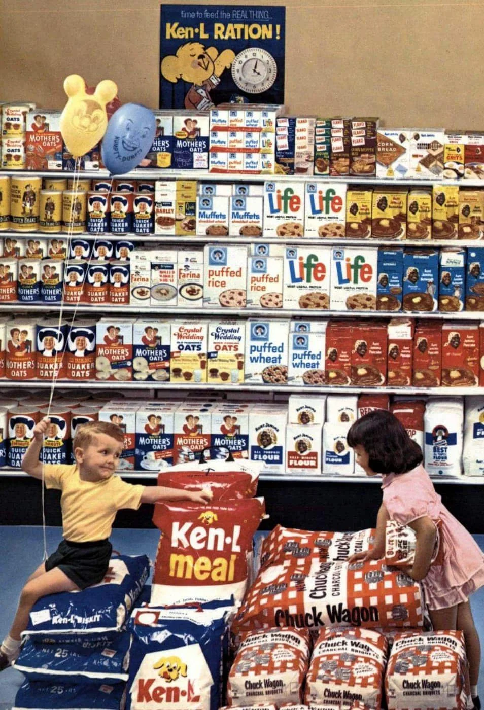 Two children in a grocery store sit on large bags of Ken-L and Chuck Wagon dog food. Behind them is a wall of colorful cereal boxes like Life, Rice Krispies, and Shredded Wheat. The boy holds balloons with cartoon faces.