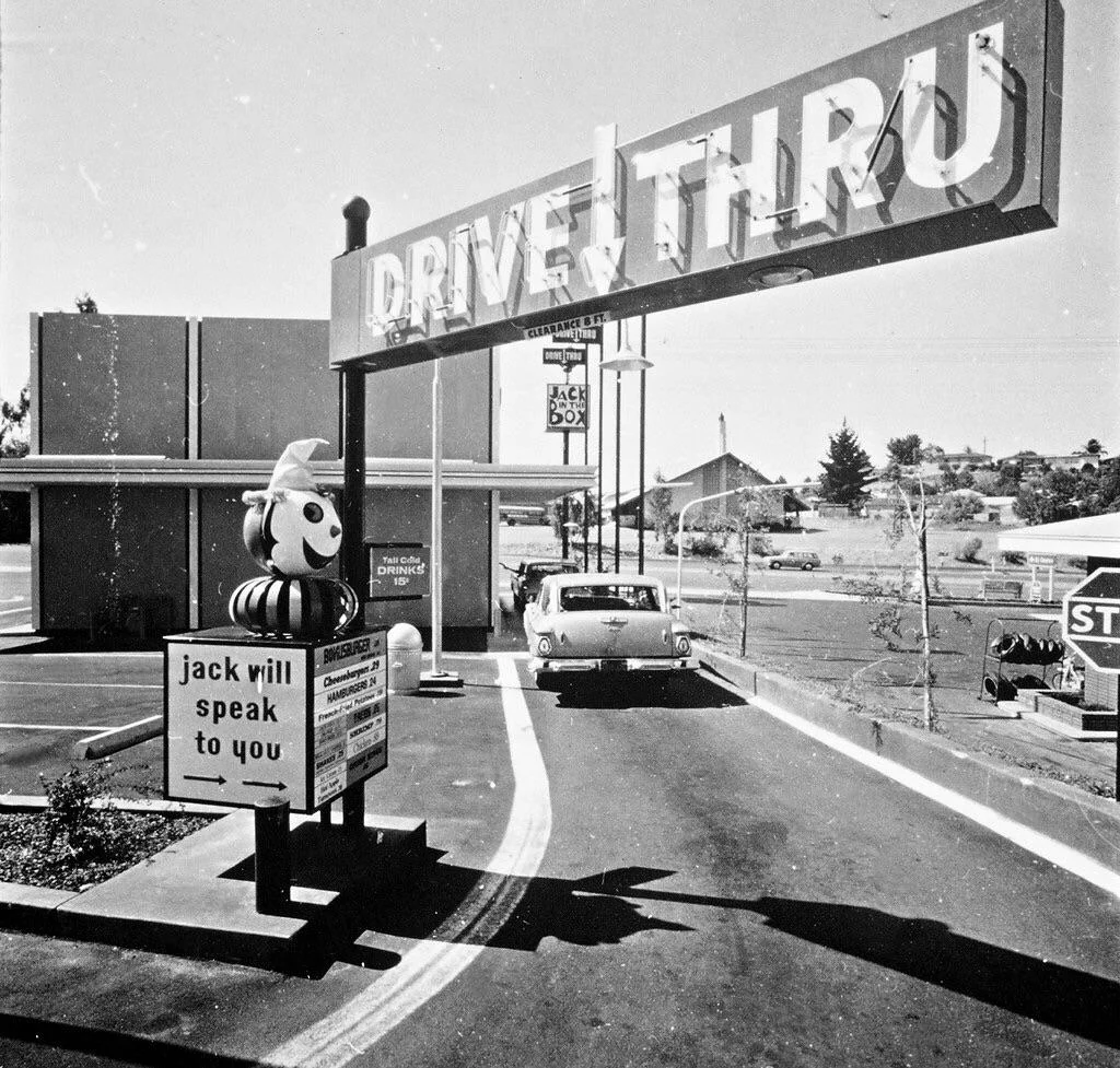 Black-and-white photo of a vintage Jack in the Box drive-thru. A car waits beside the order speaker, topped with a clown head. A large “DRIVE THRU” sign is above. The parking lot and buildings are visible in the background.