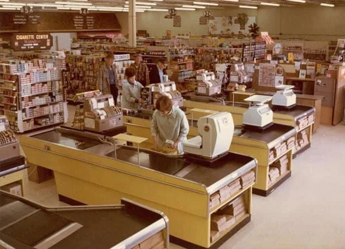 A vintage supermarket checkout area with yellow counters, old-fashioned cash registers, and paper bags. Shoppers and cashiers are visible, and shelves of products line the background.