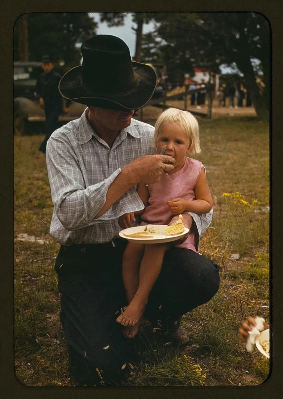 A man in a black cowboy hat feeds a young blonde girl sitting on his knee. The girl wears a pink dress and holds a plate of food outdoors, with people and tents visible in the background.