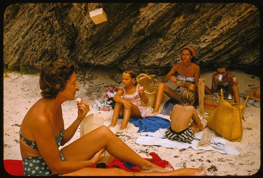 Four people, including two children, have a picnic on a sandy beach near rocky cliffs. They sit on towels, surrounded by baskets, eating and talking. The sun is shining and everyone is in swimsuits.