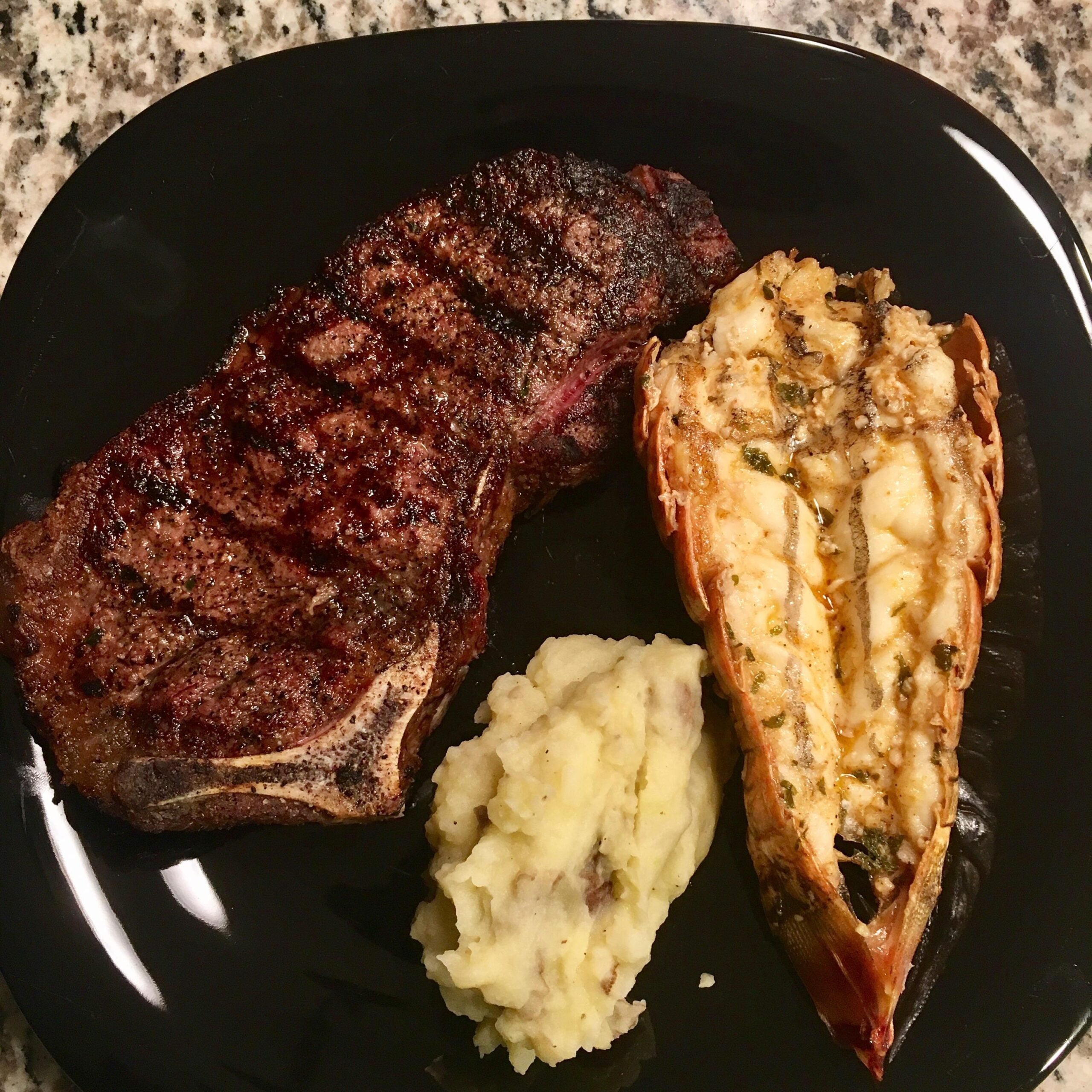 A grilled steak, baked lobster tail with herbs, and a serving of mashed potatoes are arranged on a black plate atop a granite countertop.