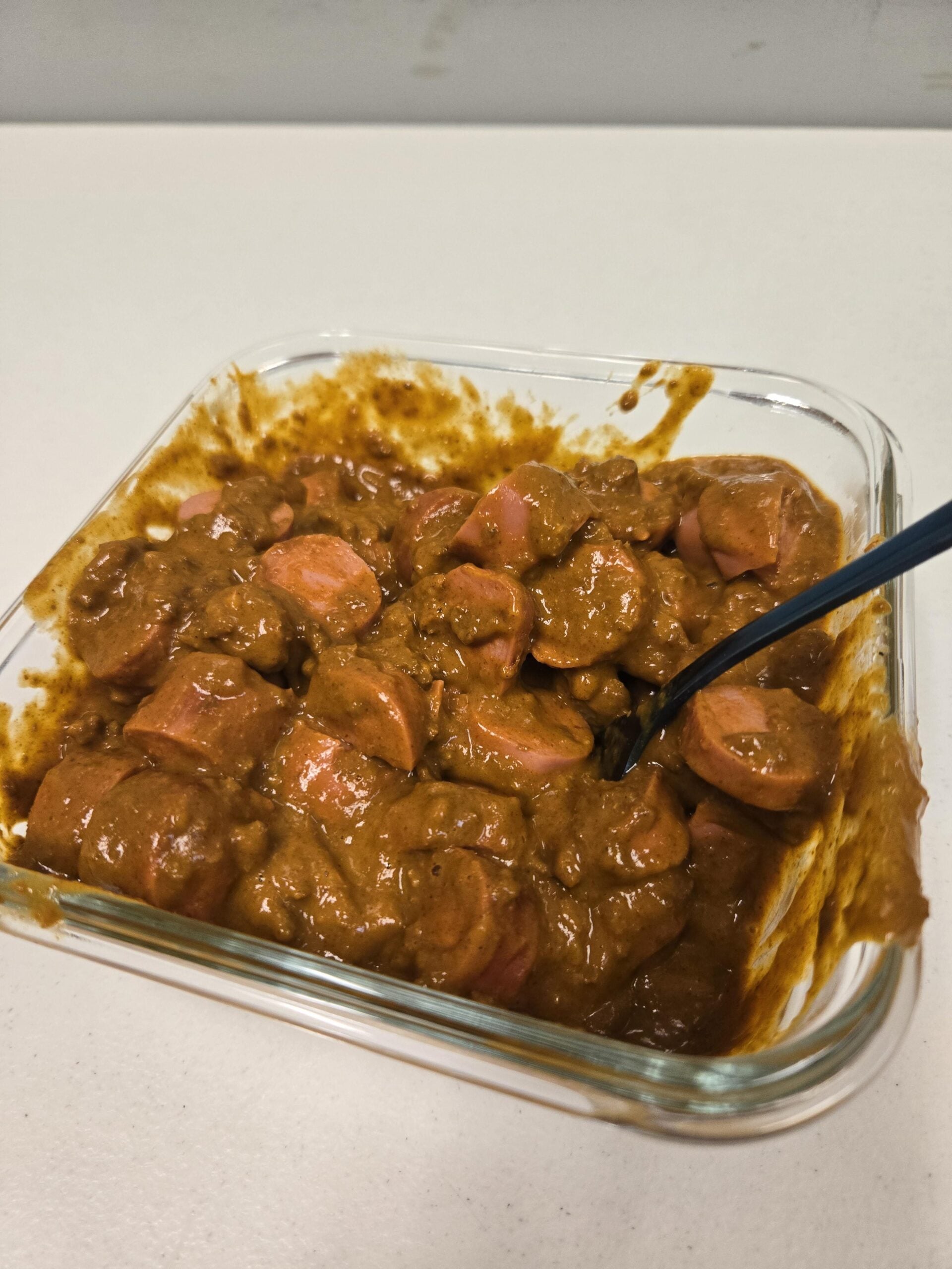 A glass container filled with sliced hot dogs mixed in a thick brown sauce, with a black plastic fork resting inside. The container is on a plain white surface.