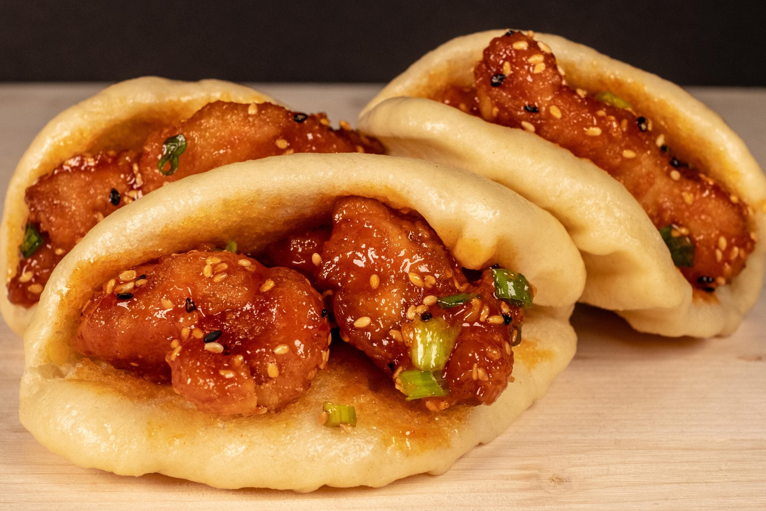 Three bao buns filled with crispy glazed chicken, garnished with sesame seeds and chopped green onions, are arranged on a light wooden surface with a dark background.