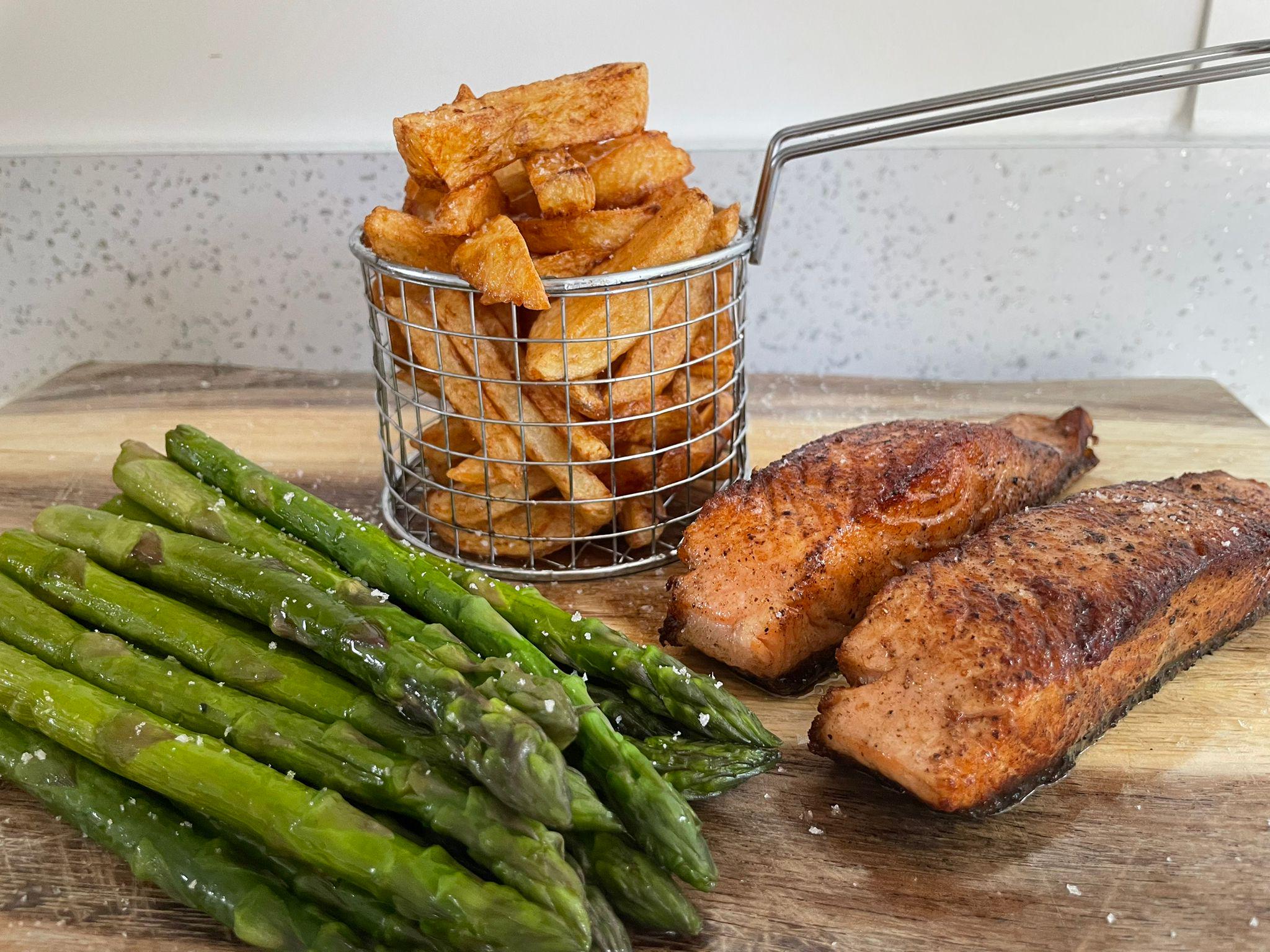 Grilled asparagus, two seasoned salmon fillets, and a metal basket filled with thick-cut fries are arranged on a wooden board.