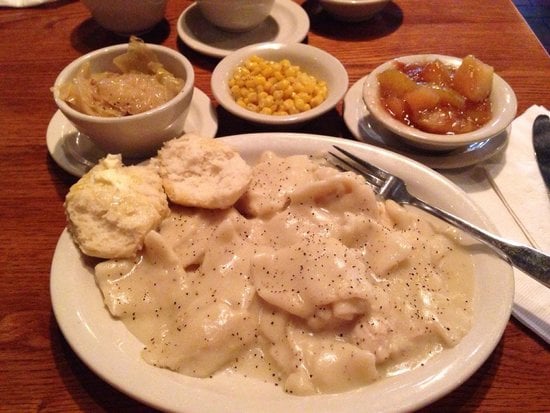 A plate of chicken and dumplings with black pepper, a biscuit, and sides of creamed corn, stewed cabbage, and glazed apples on a wooden table. A fork and napkin are placed nearby.