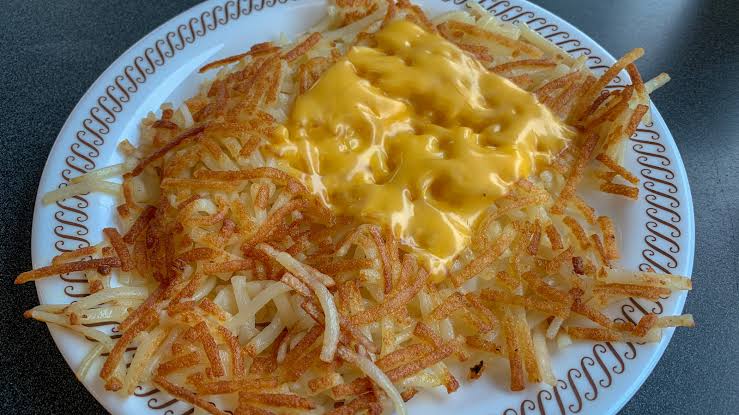 A white plate with crispy hash browns topped with melted yellow cheese, set on a dark table. The plate has a brown decorative border.
