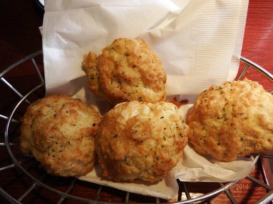 Four golden-brown biscuits with herbs are placed on a white napkin inside a metal basket on a wooden table.