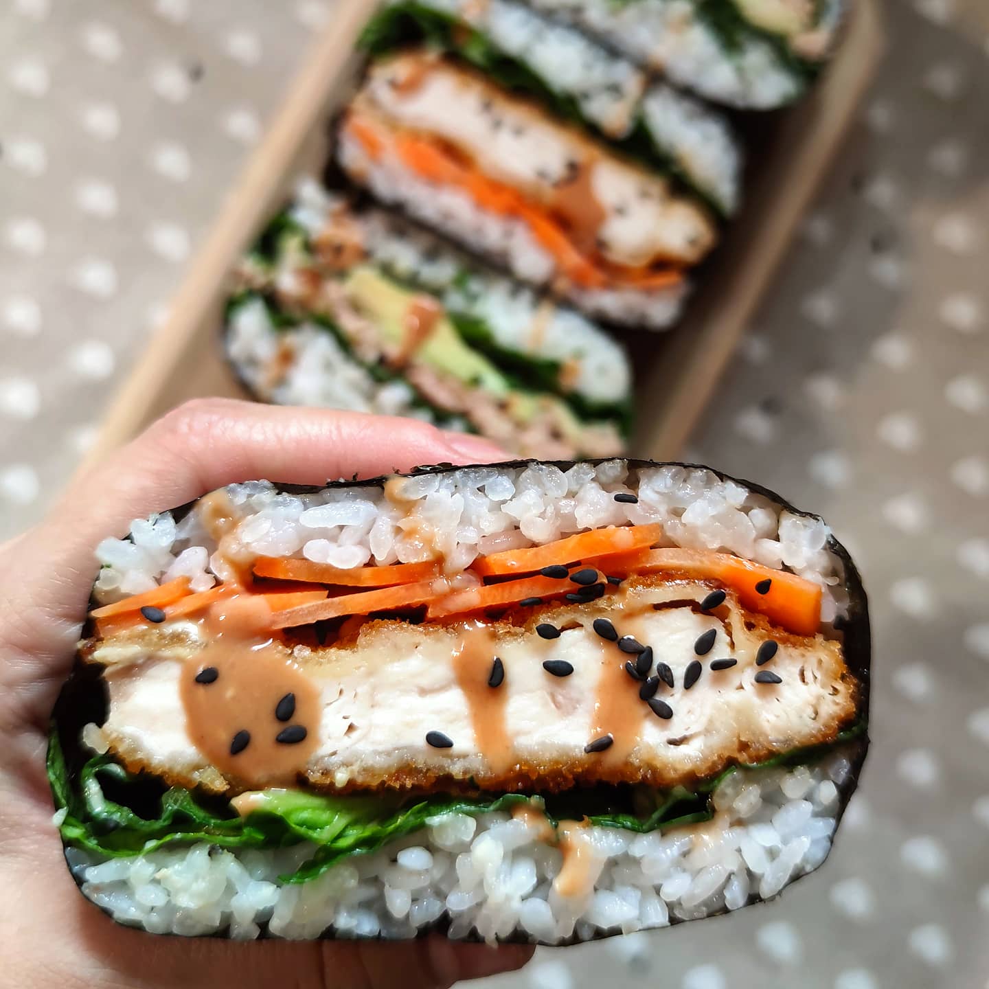 A hand holds a sushi sandwich with rice, lettuce, carrot, breaded tofu or chicken, sauce, and black sesame seeds, wrapped in seaweed. More sushi sandwiches are blurred in the background.