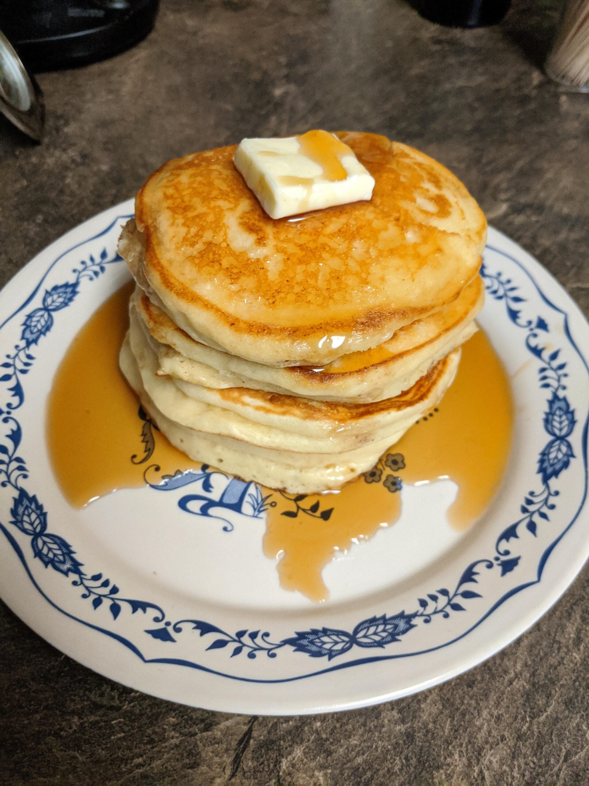 A stack of four pancakes topped with a pat of butter and maple syrup on a white plate with a blue floral pattern, sitting on a brown countertop.