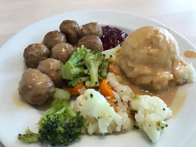 A plate with Swedish meatballs and gravy, mashed potatoes with gravy, lingonberry sauce, and steamed mixed vegetables including broccoli and cauliflower.