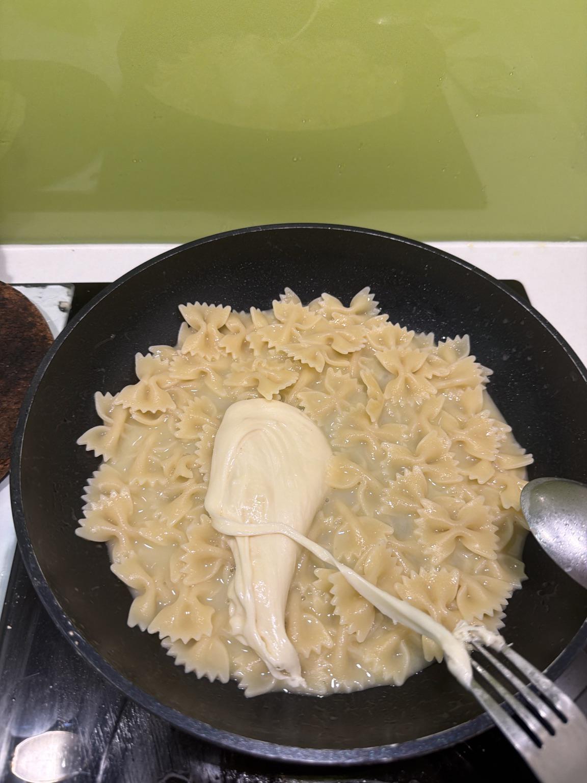 A pan of cooked farfalle pasta with a piece of melting mozzarella cheese being stretched with a fork, set on a stovetop against a green backsplash.