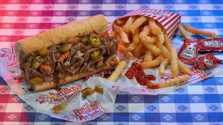 A beef sandwich with peppers, crinkle-cut fries, and ketchup packets are served on branded paper on a blue and white checkered tablecloth.