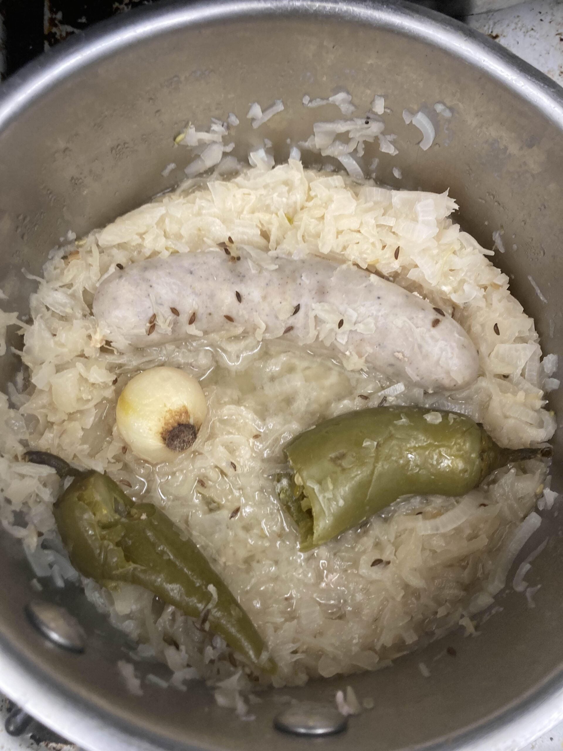 A pot filled with sauerkraut, a white sausage, a whole peeled onion, two green peppers, and visible caraway seeds simmers in liquid—teetering on the edge of one of those hilarious cooking disasters that somehow promise a truly memorable meal.