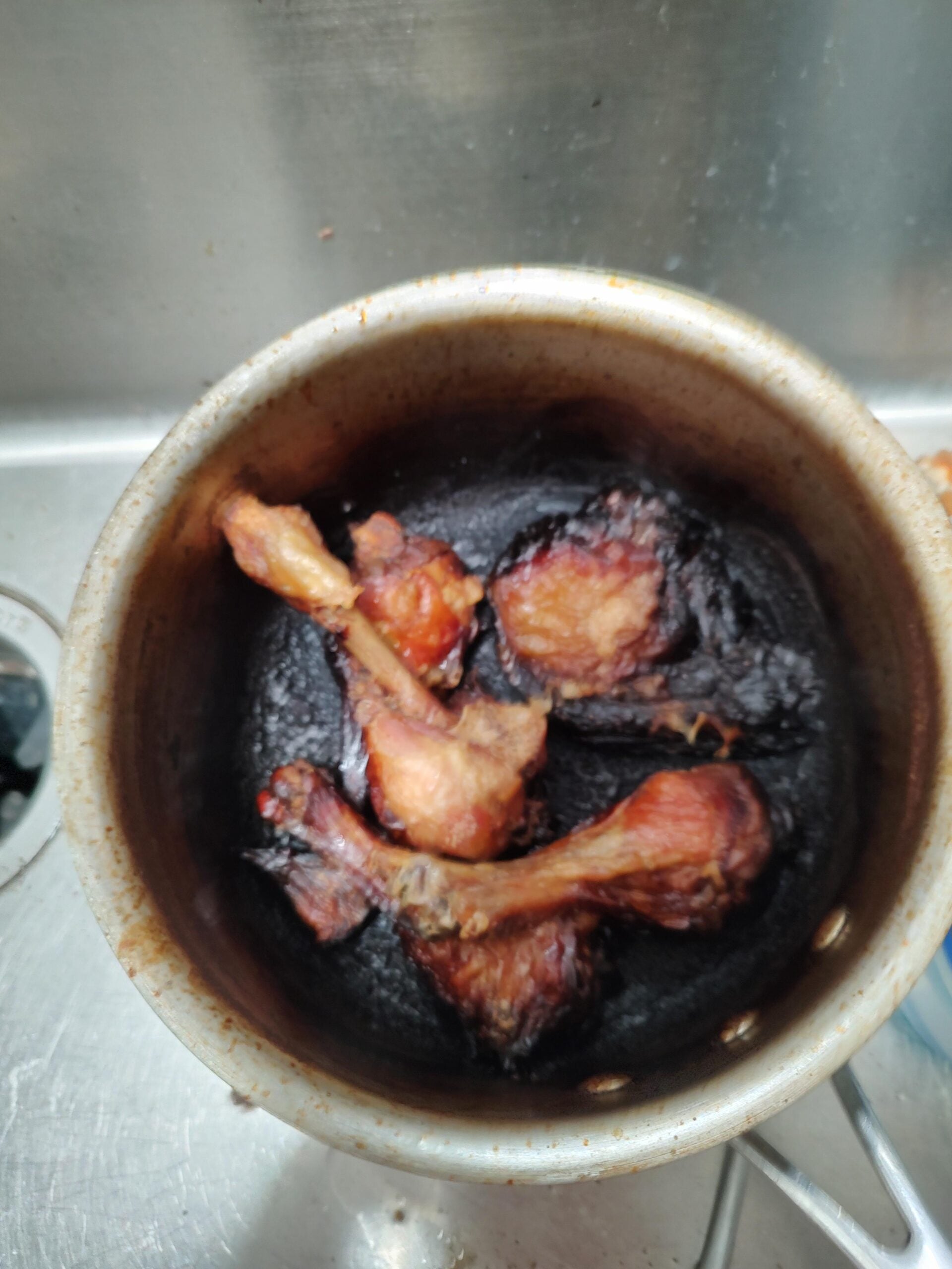 Four drumsticks are sitting on top of a thick, burnt black layer in a metal pot, showing overcooked or burnt food. The pot is placed in a kitchen sink.