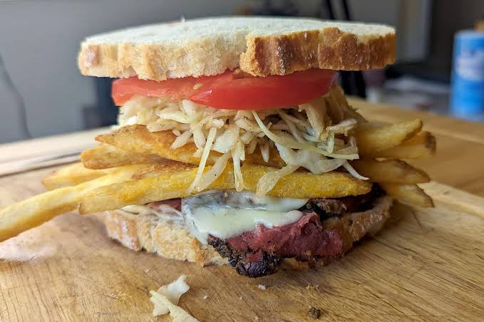 A sandwich on sliced bread filled with tomato, shredded cabbage or slaw, French fries, melted cheese, and slices of meat, all stacked on a wooden surface.