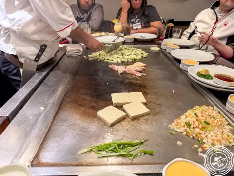 A chef prepares food on a hibachi grill, with tofu, vegetables, and chicken cooking. Diners sit around the grill, watching, with plates and sauces in front of them.