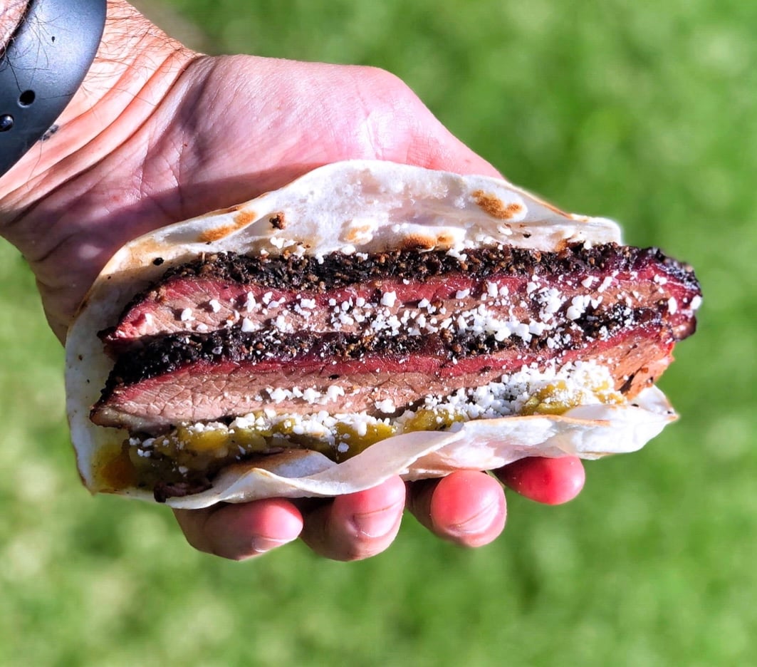 A hand holds a taco filled with thick slices of smoked brisket, sprinkled with cheese, wrapped in a soft tortilla. A blurred green lawn is visible in the background.
