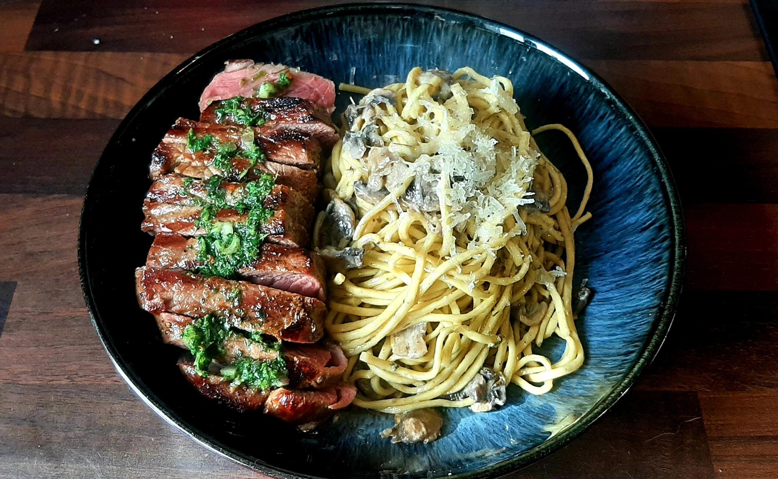 A black plate with sliced steak topped with green herbs beside a serving of spaghetti mixed with mushrooms and topped with grated cheese.