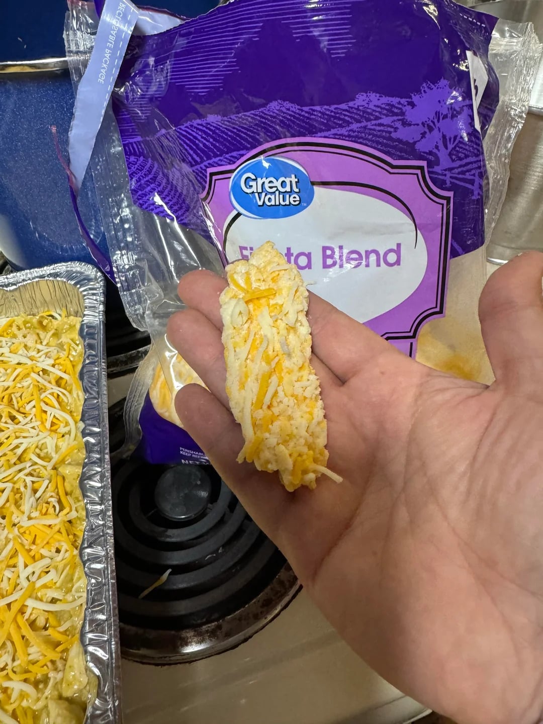 A hand holds a clump of shredded cheese pressed together. In the background, there’s an open bag of Great Value Fiesta Blend, known as one of the best shredded cheese options, and a foil tray of cheesy food on a stove.