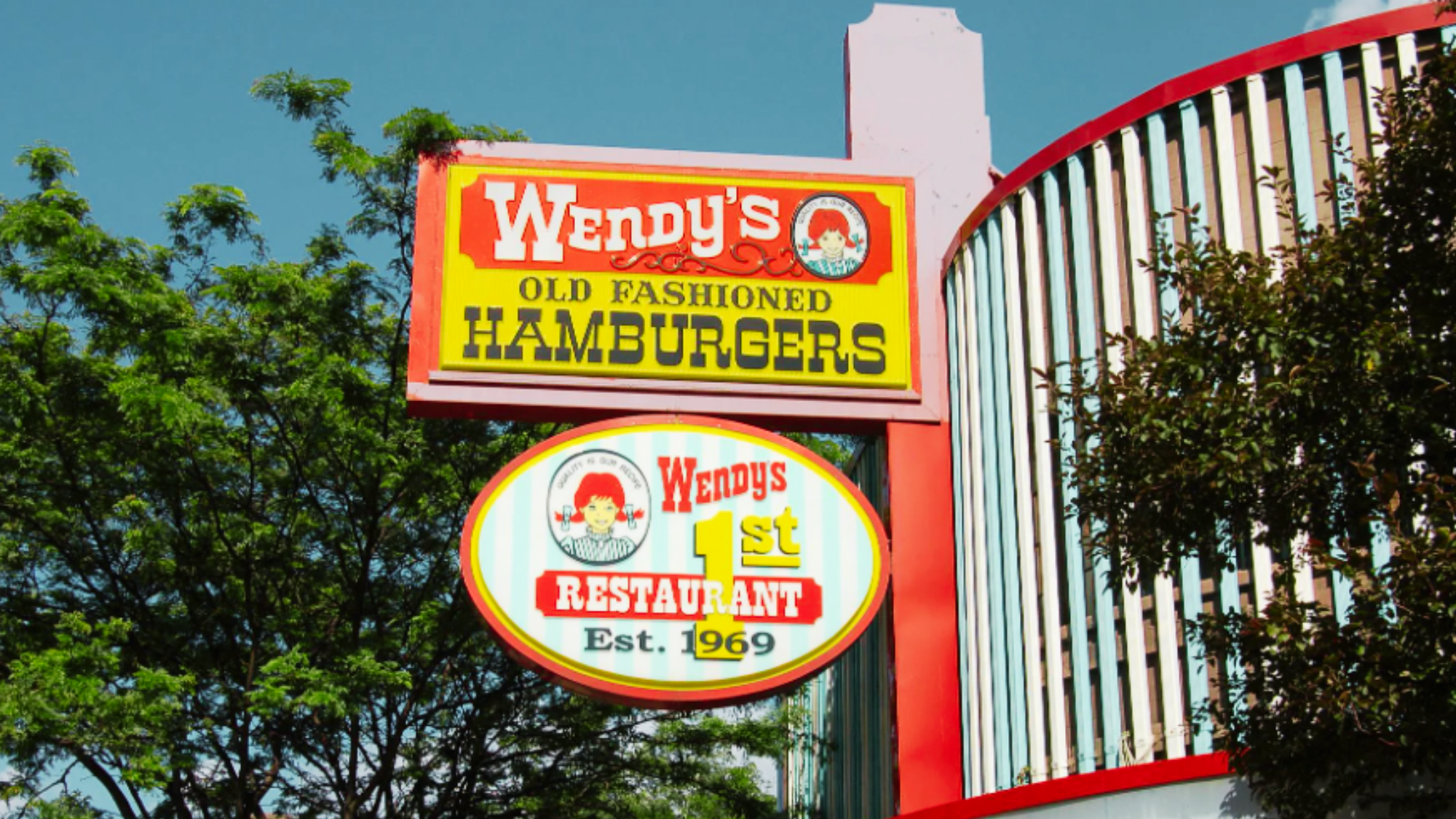 A Wendy’s sign reads “Wendy’s Old Fashioned Hamburgers” with a smaller sign below saying “Wendy’s 1st Restaurant Est. 1969,” surrounded by trees and part of a building, marking the origins of classic Wendy’s foods.