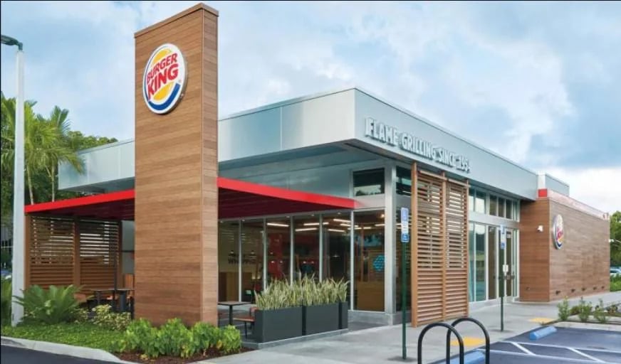 A modern Burger King restaurant with large windows, wood paneling, a tall sign featuring the Burger King logo, outdoor seating, and a bike rack in front, set on a sunny day with palm trees nearby.